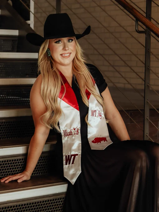 Blonde lady wearing a black felt cowgirl hat is sitting on a stairwell leaning back on one hand. She is in a black dress with a maroon and white graduation stole with WT and a buffalo on the sides. 