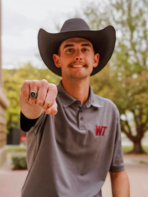 A person wearing a wide-brimmed cowboy hat and a "WT" logo polo shirt is showing a ring prominently towards the camera. There's a background of trees and a building.