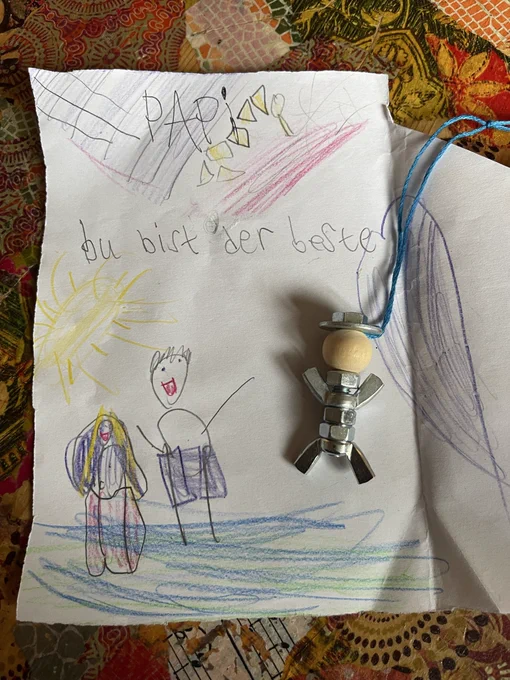 Handmade children’s drawing on white paper featuring colorful crayon sketches and a small homemade angel ornament attached with blue string. At the top, the word “PAPA” is written in large childlike letters with decorative shapes and scribbles around it. Beneath it, handwritten German text reads “du bict der beste,” intended to mean “du bist der beste” (“you are the best”).

The drawing includes a bright yellow sun in the upper left corner, blue and green scribbles representing ground or water, and two smiling stick-figure people standing together near the bottom. One figure has long yellow hair and colorful clothing, while the other has short dark hair and purple clothing.

Attached to the paper is a small angel-like craft made from metal nuts and bolts, wooden beads, and blue string, giving the artwork a handmade gift-like appearance. The paper rests on a richly patterned, multicolored tablecloth with floral and mosaic-style designs.