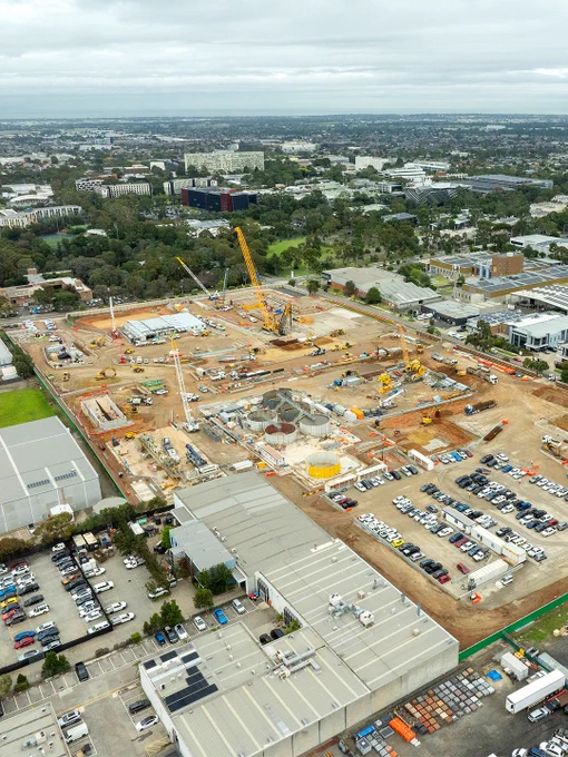 Aerial view of a construction site with multiple cranes, surrounded by urban buildings and a parking lot. The construction area appears busy, with various materials and equipment visible.
