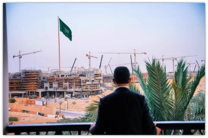 Anies Baswedan berdiri di balkon sambil memandang kawasan pembangunan Ad Diriyah di pinggiran Kota Riyadh yang tengah berkembang pesat.

Dari belakang terlihat ia mengenakan setelan jas gelap menghadap hamparan proyek konstruksi besar dengan deretan crane, bangunan yang sedang dibangun, serta bendera Arab Saudi yang berkibar tinggi di tengah area proyek.

Lanskap gurun, pepohonan palem, dan aktivitas pembangunan modern menggambarkan transformasi Ad Diriyah sebagai salah satu kawasan strategis dalam pengembangan Riyadh masa depan.