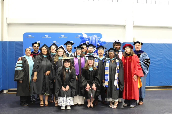 A group of graduates and faculty members from the University at Buffalo pose for a photo in their graduation gowns and regalia in front of a blue backdrop with the university logo. 