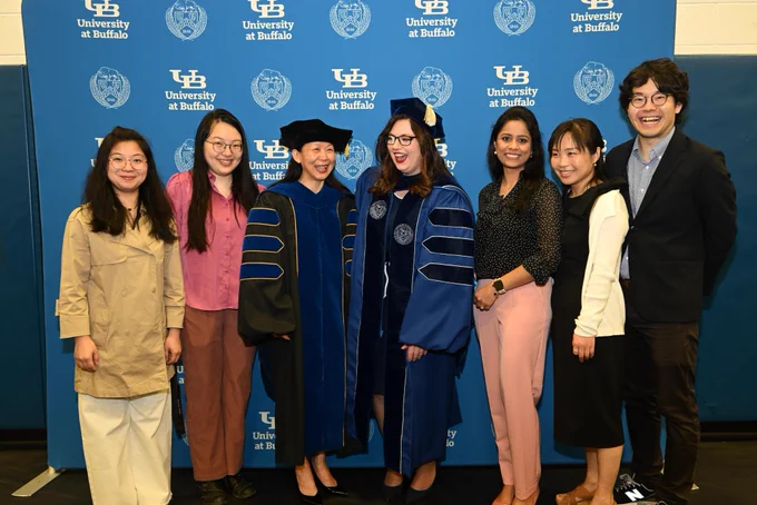 A group of eight people stand smiling in front of a blue backdrop with the \"University at Buffalo\" logo. Two are in graduation gowns; others are in business casual attire, creating a celebratory and academic atmosphere.