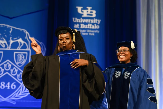 Two smiling women in graduation gowns stand before a University at Buffalo backdrop. One woman puckers her lips and gestures with her finger. The other wears glasses and a graduation cap with a white tassel.