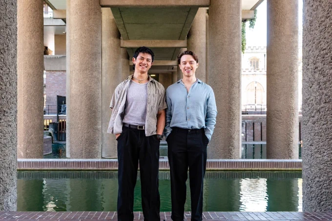 A photo showing two young men stood amongst brutalist architecture, smiling to the camera 