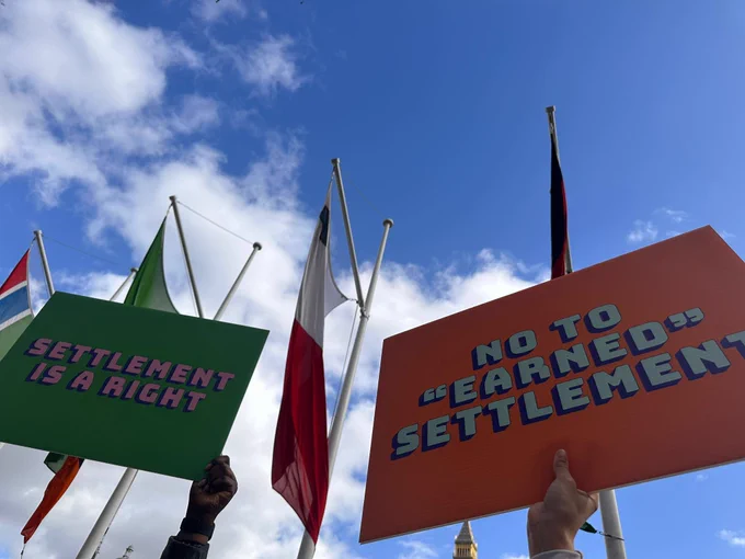 Two placards against a blue sky with a range of national flags say "settlement is a right" and "no to earned settlement"