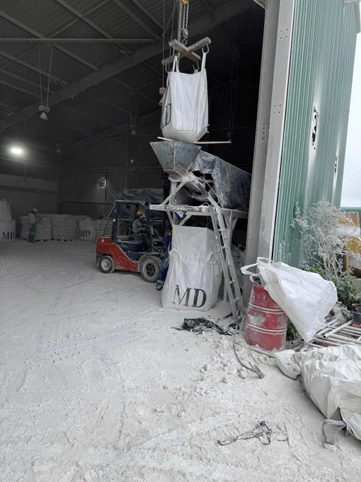 The image shows the interior of an industrial warehouse with a forklift lifting a large white bag into a funnel system. Numerous other white bags labeled "MD" are stacked in the background.