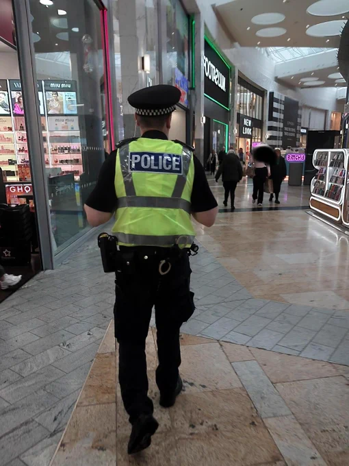 A police officer in uniform walks through a shopping centre, wearing a high-visibility vest.
