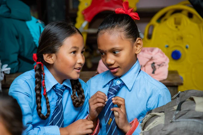 Two school children talking to each other as they sit close. One of the children is playing with their school tie. 