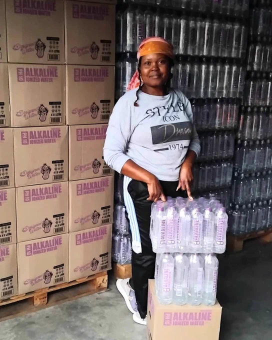 Grancious, smiling, standing next to stacks of Designer Water ph10 in a warehouse.