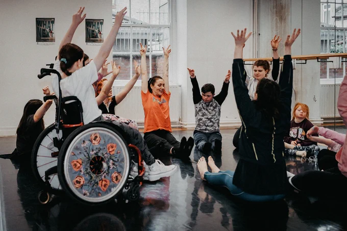 Photo of a group of adults and children in a dance studio, all with their hands up. One child is using a wheelchair. 