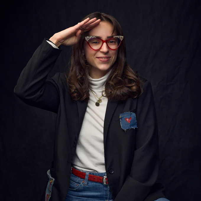 A person wearing red glasses and a black blazer with a heart patch, saluting in front of a dark, textured background.