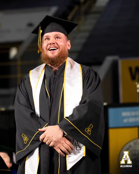 Country music artist and App State alumnus Luke Combs smiles while standing on stage during Appalachian State University’s commencement ceremony inside Holmes Convocation Center. He wears a black graduation cap and gown with a white stole trimmed in gold.