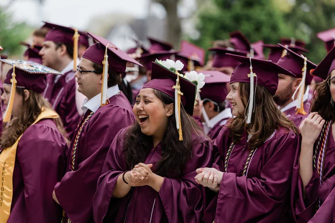 A group of graduates in maroon caps and gowns stands together, celebrating. One person in the foreground is clapping and smiling, and there are decorative flowers on their cap. Trees and a building are visible in the background.