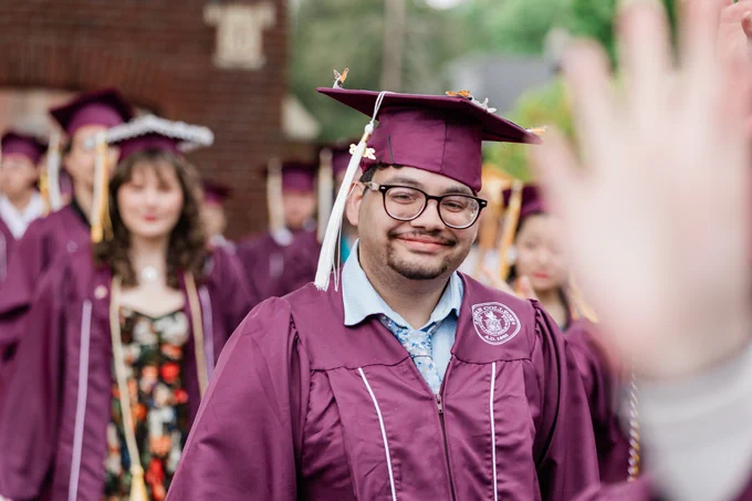 A person in a maroon graduation gown and cap is smiling and standing in a procession. Others in similar attire are in the background. A hand is partially visible in the foreground.