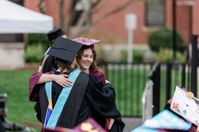 Two graduates in caps and gowns embrace at a graduation ceremony. One wears a decorated maroon cap, and the other has a sash with blue and yellow stripes. A brick building and greenery are in the background.