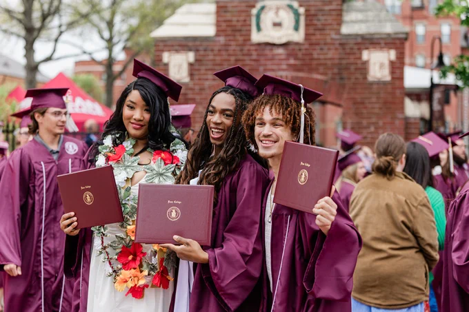 Three graduates in maroon caps and gowns hold diplomas at a ceremony. The scene is lively, with people in the background and a brick structure visible.
