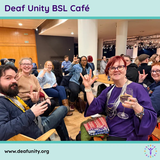 Smiling group at Deaf Unity BSL Café sitting around tables, holding drinks and showing sign-language handshapes.