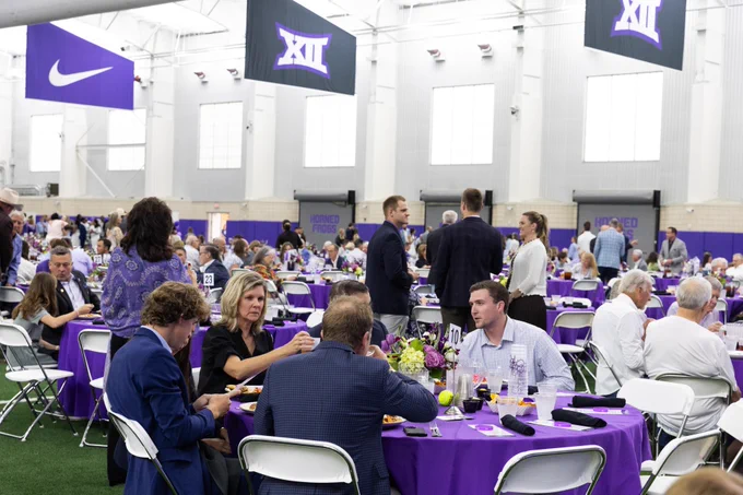 People are gathered in TCU's Indoor Football Practice Facility with purple tablecloths, where various groups are dining. Overhead banners display the Nike logo and the Big 12 Conference logo. The setting appears festive and social.