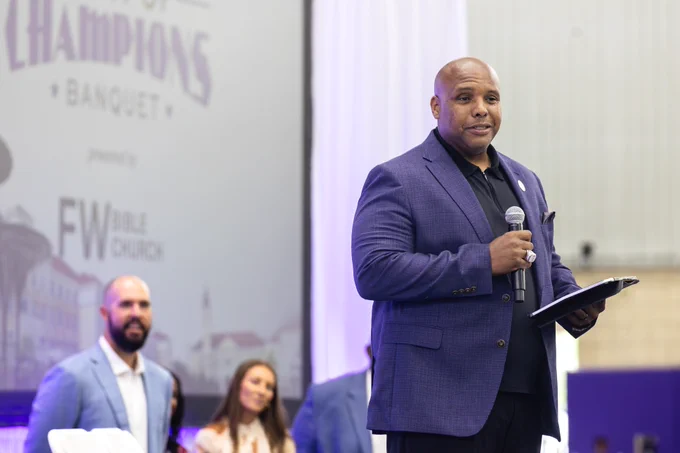 A person in a purple blazer speaks while holding a microphone and a folder, standing on a stage next to a FW Bible Church sign. Other people are visible in the background near a large display screen.
