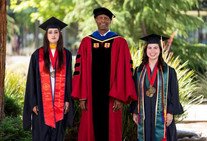 Three individuals in academic regalia stand outdoors. Two are wearing black graduation caps and gowns with colorful sashes and medals. The third, centered, is in a red and black robe with a doctoral cap. They are surrounded by greenery.
