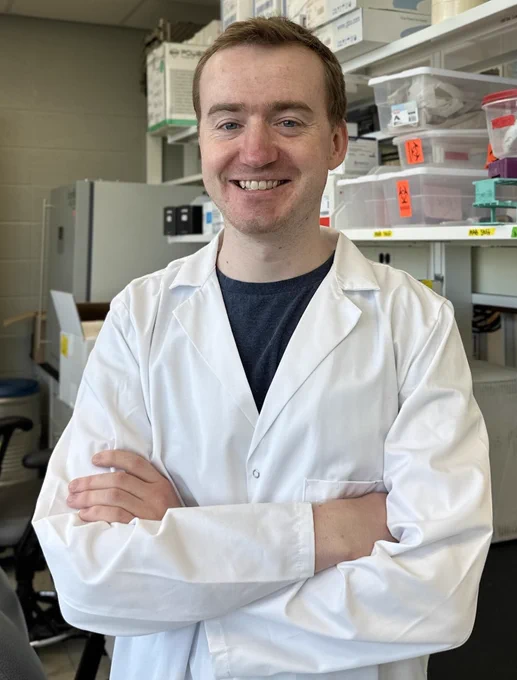 Jared Godfrey smiles at the camera. He's in a lab and is wearing his lab coat. 