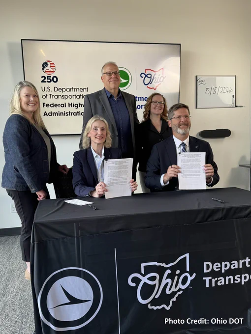 Five individuals are gathered for a formal event at a table with an "Ohio Department of Transportation" banner. Two seated people hold signed documents. FHWA and ODOT logos on a large monitor behind them. Smaller white board beside the monitor showing 5/8/2026. Photo Credit: Ohio DOT.