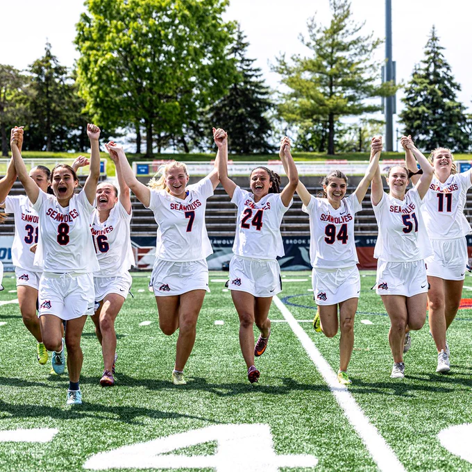 Women’s lacrosse players celebrate on the field with raised arms after a game.