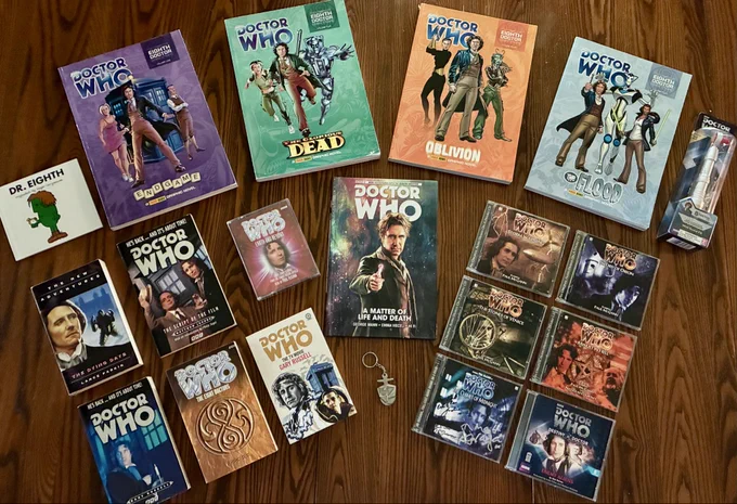 A collection of 8th Doctor themed books, comics, and audios, arranged on a wooden table.