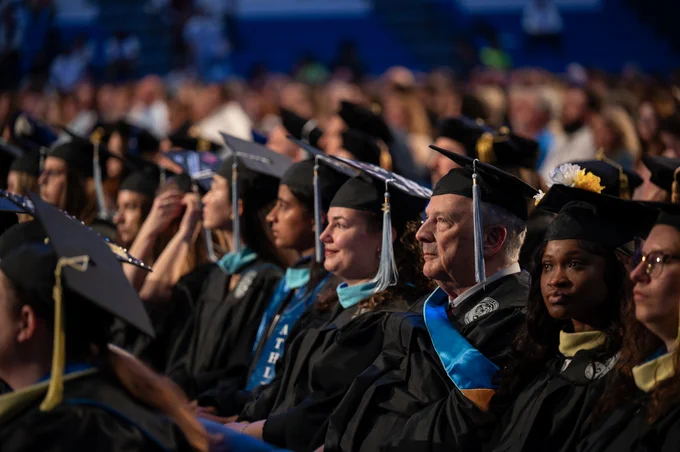 A diverse group of graduates in black gowns and caps sit in rows at a graduation ceremony. The caps have tassels and some are decorated with flowers. The gowns have light blue sashes.