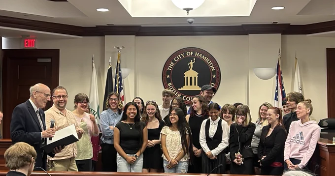 Hamilton Mayor Moeller stands at a podium in City Council chambers presenting a certificate and proclamation to Director Lischak and members of the Hamilton High School Varsity Winter Guard. The students are smiling and gathered in front of the City of Hamilton, Butler County, Ohio seal following their historic Top 20 finish at the WGI World Championships.