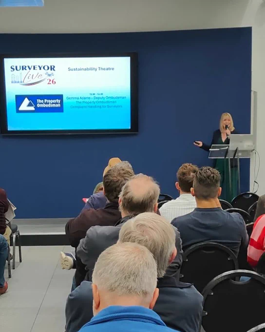 A smartly dressed woman stands at the front of a room presenting to an audience. She is speaking into a microphone and pointing towards a presentation screen on the wall.