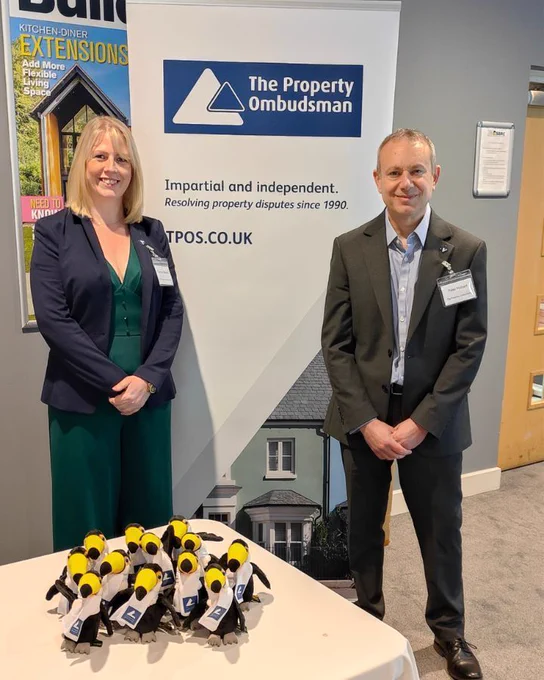 A smartly dressed man and woman stand in front of a roller banner promoting The Property Ombudsman at an industry event. A group of branded toucan toys sits on the table in front of them.