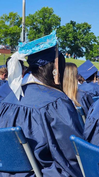 A side view of a grad wearing a grad cap decorated with blue butterflies and a white bow. There is a small photo frame hanging from the tassel with a childhood photo of the grad and a loved one.