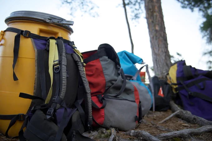 A yellow plastic barrel and several colorful backpacks and bags are placed on the ground in a forested area. Trees and a slightly overcast sky are visible in the background.