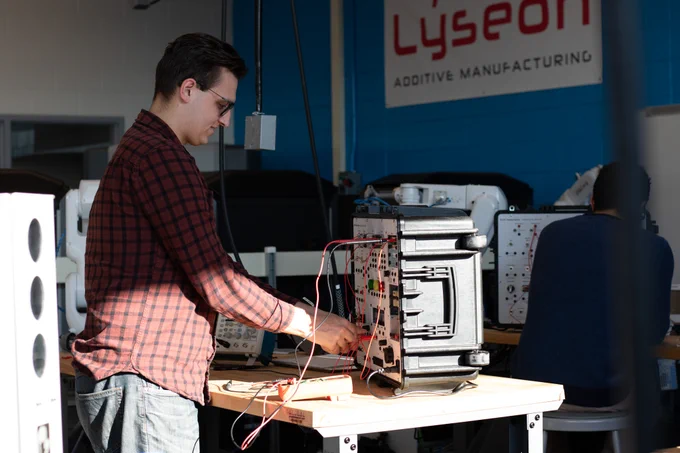 Technician adjusting wires on a complex electronic device at a workstation in a lab with Lyseon Additive Manufacturing signage.