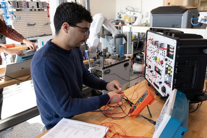 Person testing electronic circuits with multimeter probes connected to a device in a workshop filled with technical equipment.
