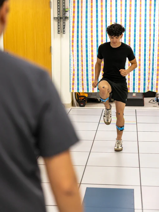 Young man wearing motion sensors performs a knee lift exercise in a motion analysis lab with a person observing him.