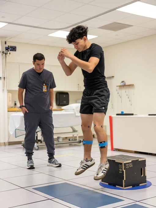 Young man wearing sensors on legs performs a jump test in a motion analysis lab under supervision of a healthcare professional.