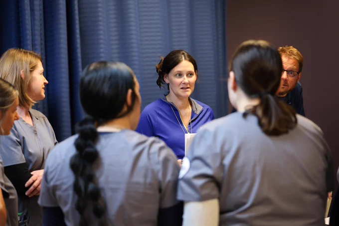 Group of nursing students in scrubs gathered in a circle in front of an instructor, engaged in a focused discussion.