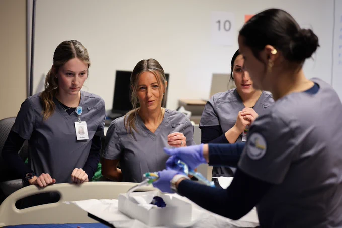 Four nursing students in scrubs gathered around a hospital bed, focusing on a medical training task.