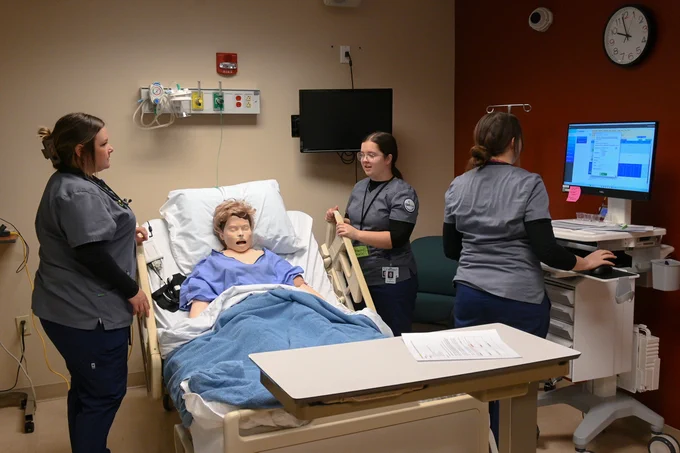 Manikin in hospital bed covered with a blue blanket, attended by three nursing students in gray scrubs during a simulation.