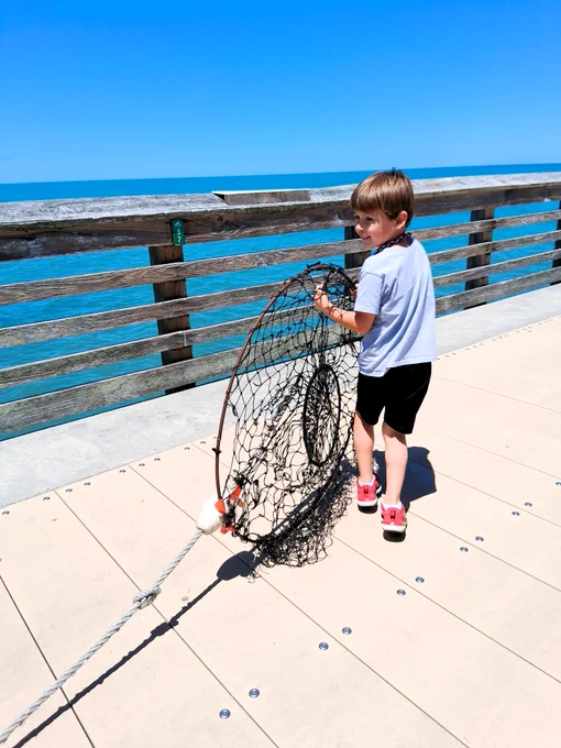 A young angler walks on a sunny pier, holding a large net. The ocean is visible in the background.