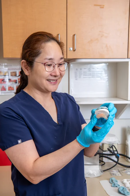 A woman wearing glasses and blue gloves holds a small container while smiling in a lab setting. She is dressed in a navy blue scrubs top, with laboratory equipment visible on the table behind her.