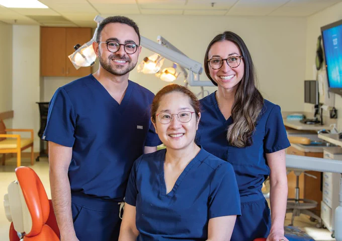 Three dental professionals stand together in a clinic. Two are positioned behind the third, who is seated in a dental chair. They are all wearing navy scrubs and smiling, with dental equipment and workstations visible in the background.
