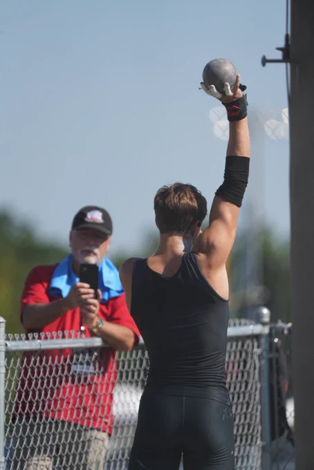 a male athlete holding a shotput above his head while his coach videos him