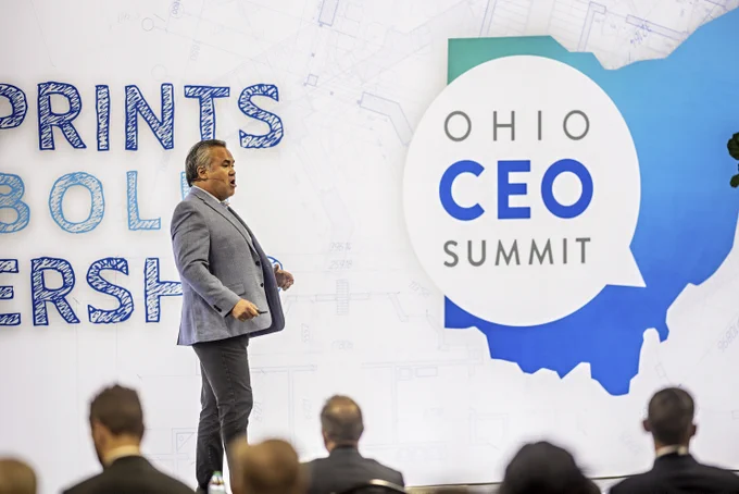 A speaker in front of an audience at the Ohio CEO Summit. The backdrop displays the event name with decorative text and design elements.