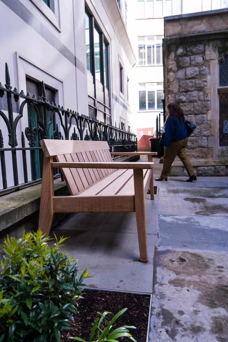 Wooden bench beside an iron fence in a narrow courtyard, with plants in front and a person walking past a stone building.