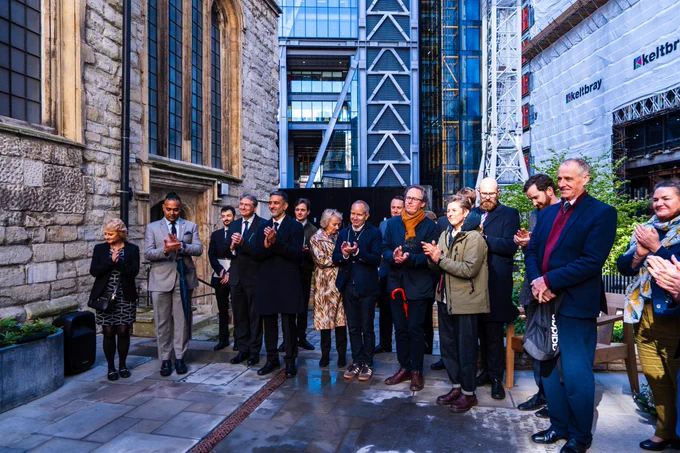 Group of people applauding in a courtyard between a historic stone church and modern glass buildings under construction.