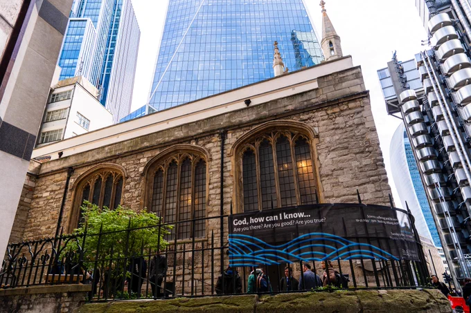 Historic stone church with Gothic windows dwarfed by modern glass skyscrapers, with people gathered behind a black iron fence.